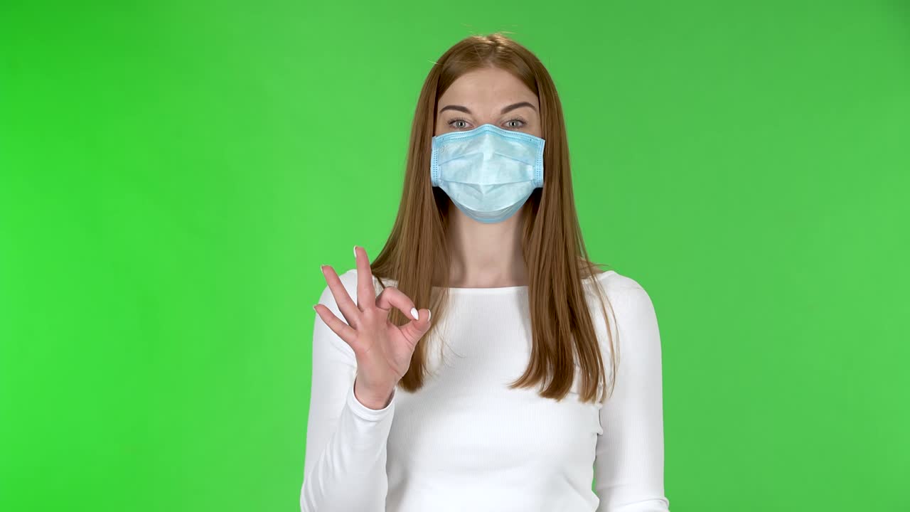 Portrait of happy pretty young girl in medical protective face mask looking at camera and showing ok sign with both hands
