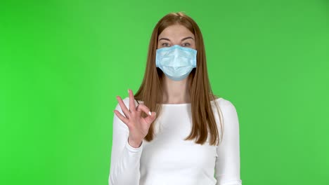 portrait of happy pretty young girl in medical protective face mask looking at camera and showing ok sign with both hands