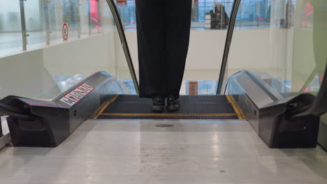 back view of woman in black pants stepping onto descending escalator with visible warning signs on escalator sides, interior of vibrant modern mall in background with people seen through glass panels