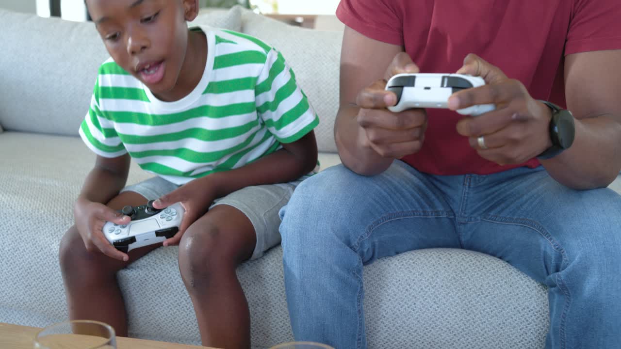 African American father and boy on sofa after game start gaming and reacting with white controllers
