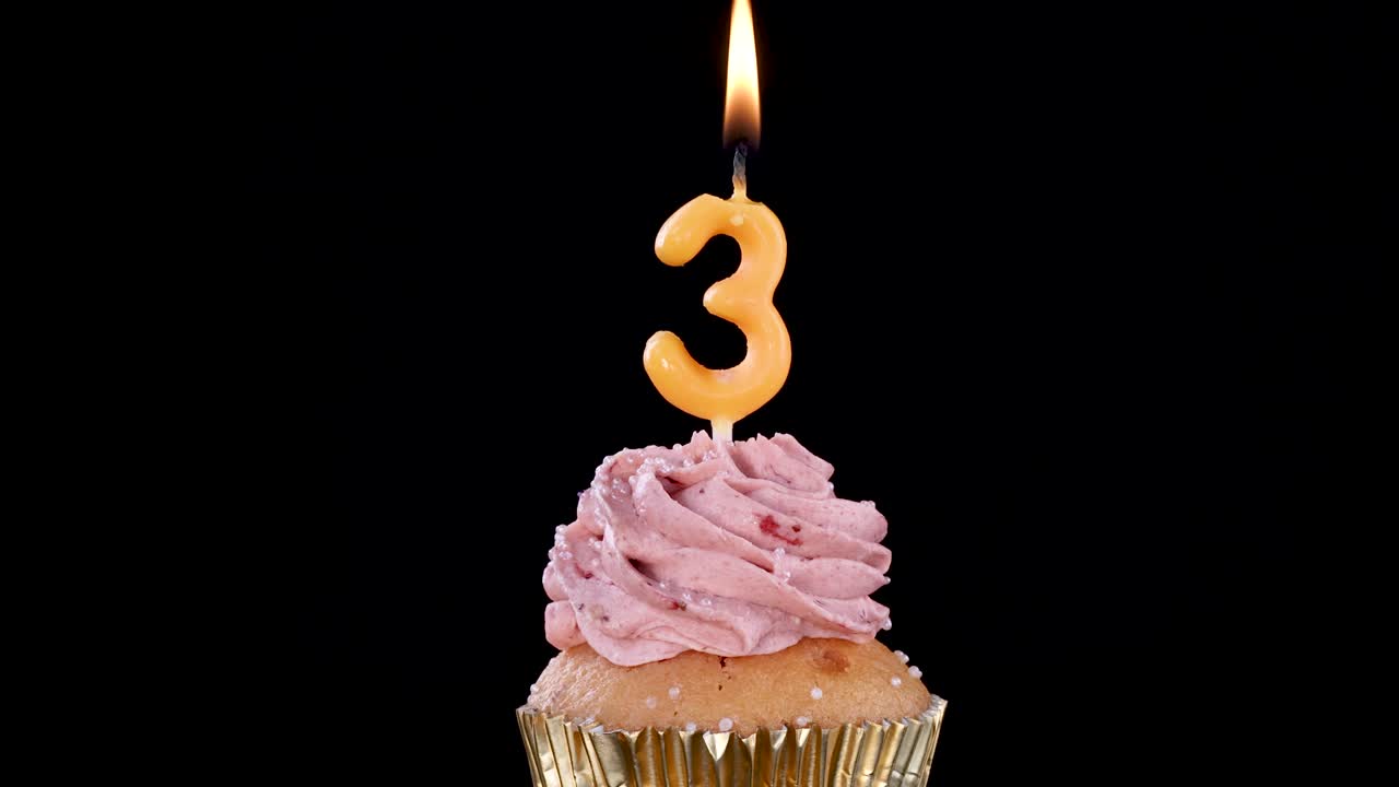 A single cupcake topped with pink frosting and a lit number three candle, shown against a black background with steady camera and bright lighting