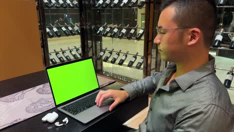 Wealthy-Asian-Millennial-Man-Scrolling-on-Laptop-with-Green-Screen-While-Grinning-Slightly,-Sitting-Inside-Luxurious-Home