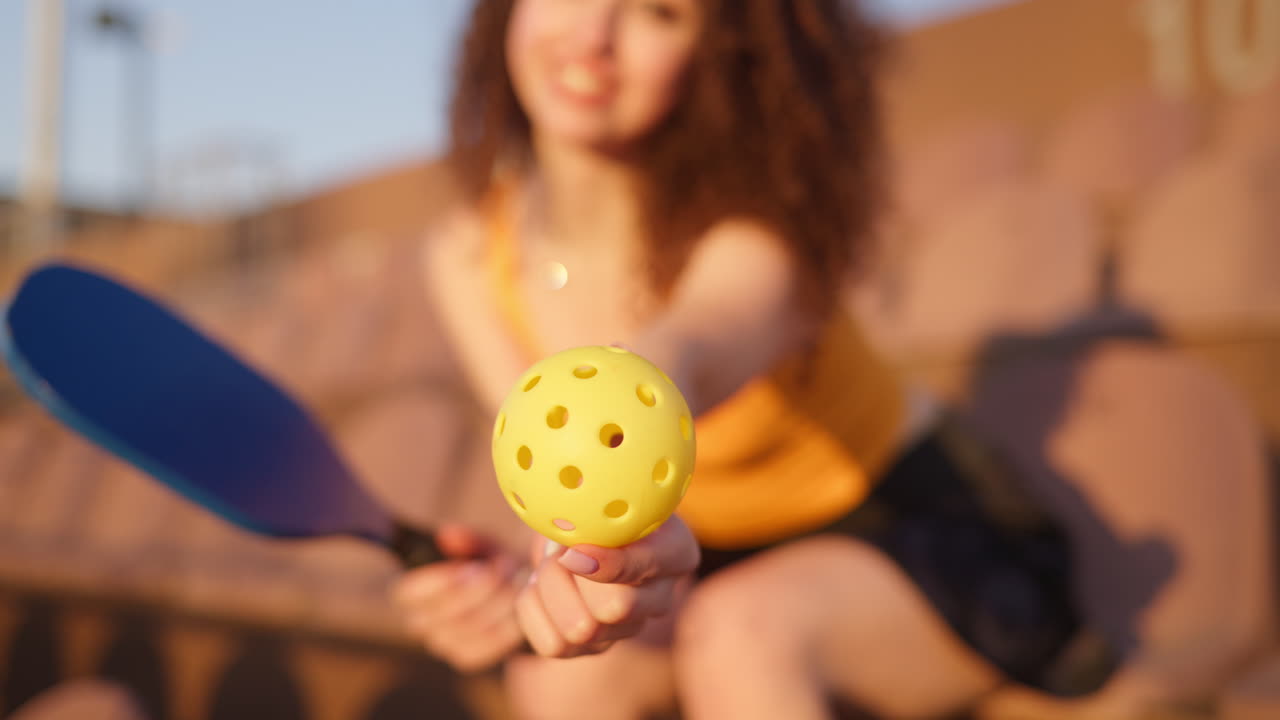 Close up of a yellow ball with a blurry view of a woman with curly hair sitting on a courtside seat, holding a pickleball racket