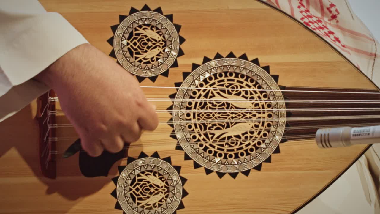 Eine detaillierte Nahaufnahme der Hand eines Musikers, der die Saiten einer traditionellen saudi-arabischen Oud zupft. Das Holzinstrument weist aufwendige dekorative Rosetten und Handwerkskunst auf
