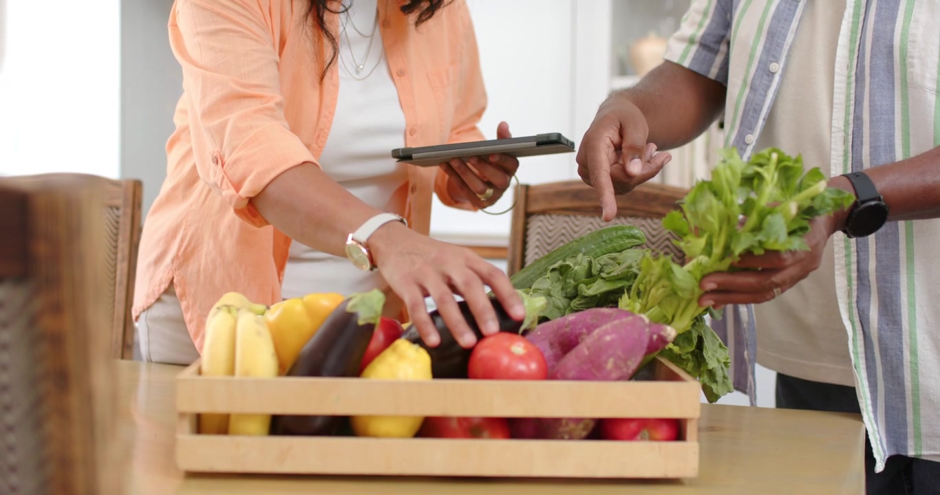 After crate arrival, senior couple unpacking and inspecting produce at kitchen table with tablet