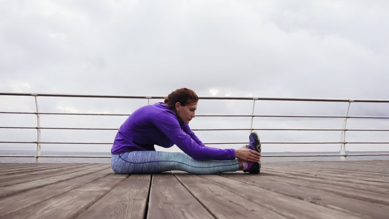 mujer atlética estirando las piernas sentada en la plataforma en la playa junto al mar temprano en la mañana. entrenamiento junto a la playa