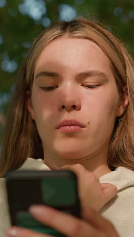 young woman in casual hoodie focuses intently on her phone while holding her hood collar, face bathed in sunlight filtering through leaves, background blurred with lush greenery