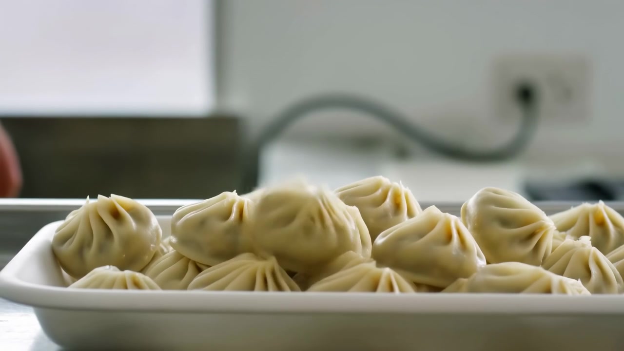 Low angle close view shows handmade pleated dumplings being transferred from metal rack into white takeout tray, with several pieces suspended above surface and fresh texture visible