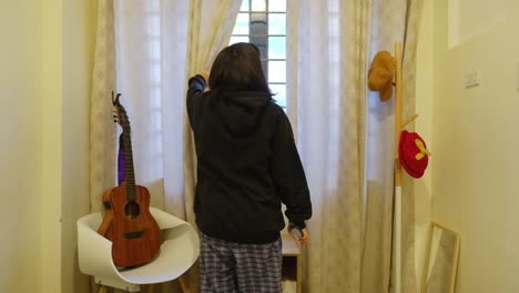 static back view of girl with black hair in jacket standing up and opening curtains in bedroom