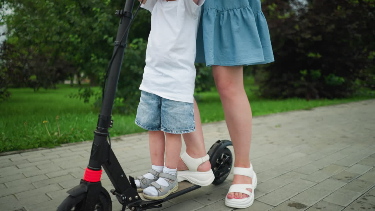 A mother and her young son share a scooter ride along a tree-lined walkway, the mother places her leg on the back tire as they move