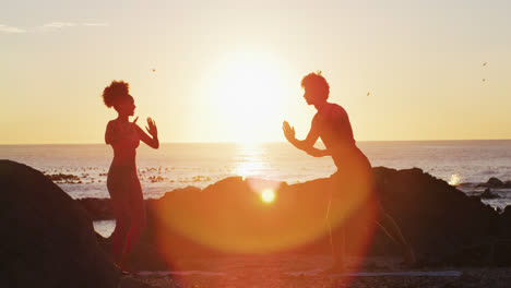 african american couple practicing yoga together on the rocks near the sea during sunset