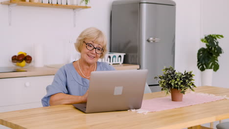 happy senior woman sitting at table in kitchen talking on video call on modern laptop