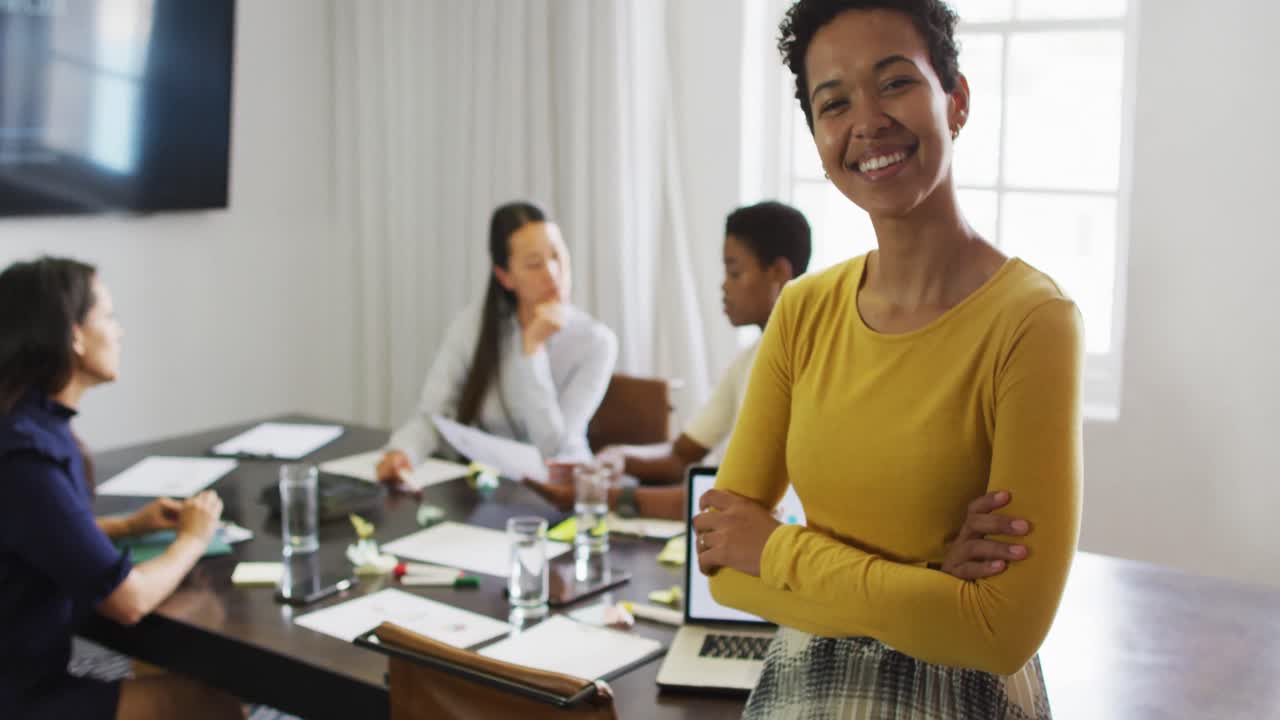 feliz mujer de negocios biracial de pie en la oficina y mirando a la cámara con colegas hablando detrás