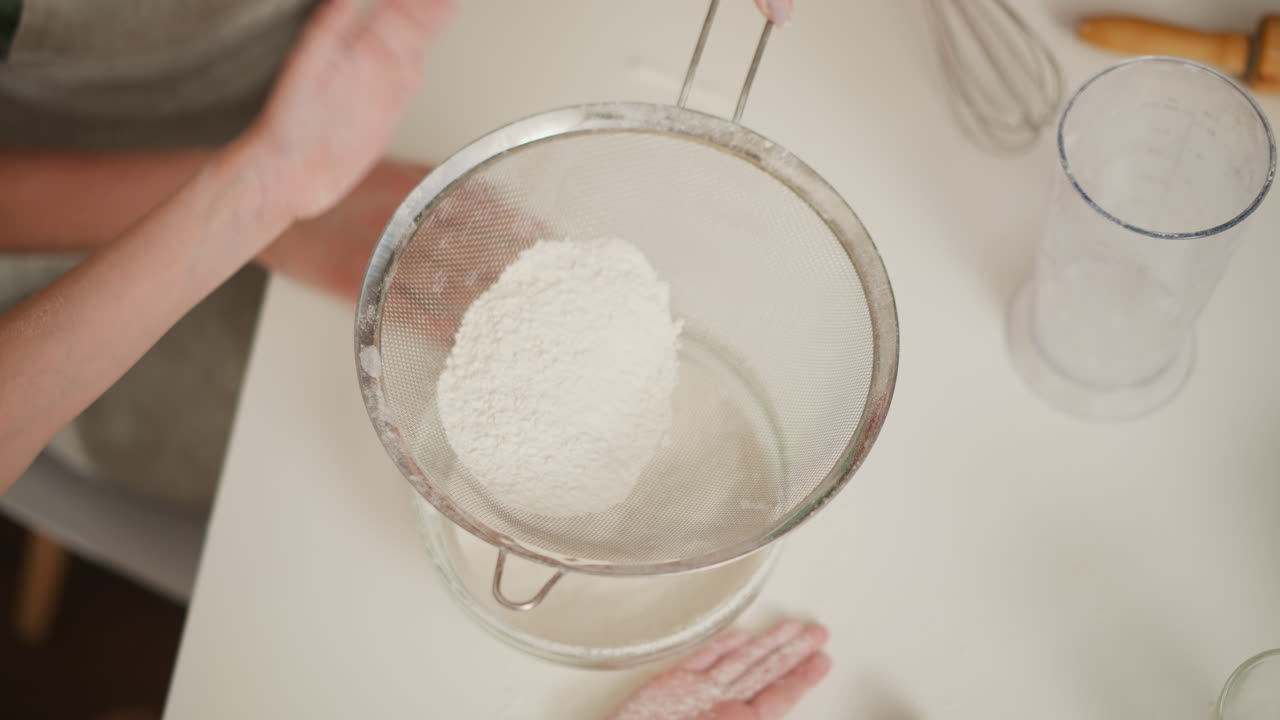 top down view of child tapping sieve filled with flour using left hand while another person firmly holds handle, flour falling into clear glass bowl on white kitchen counter