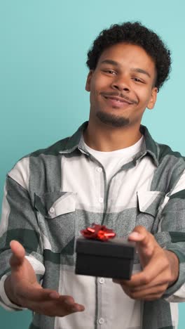 happy man giving gift box and looking at camera in blue studio