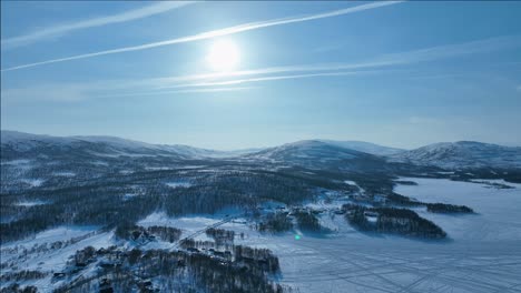 backward aerial video above snowy mountains down to village and lake surface with small island, sunny winter day in northern sweden at norwegian border, joesjo