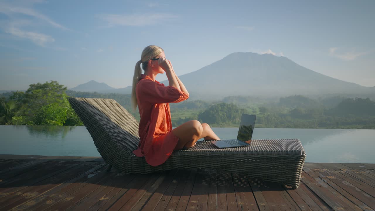 Confident blond business woman in bathrobe working on laptop whilst enjoying view of Mount Agung at pool