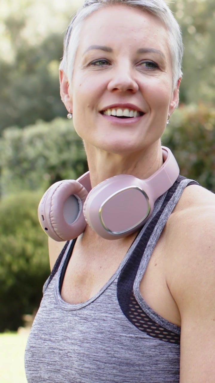 Video verticale: Panoramica che rivela una donna che parla sorridendo al parco dopo una corsa, con cuffie rosa e canotta grigia.