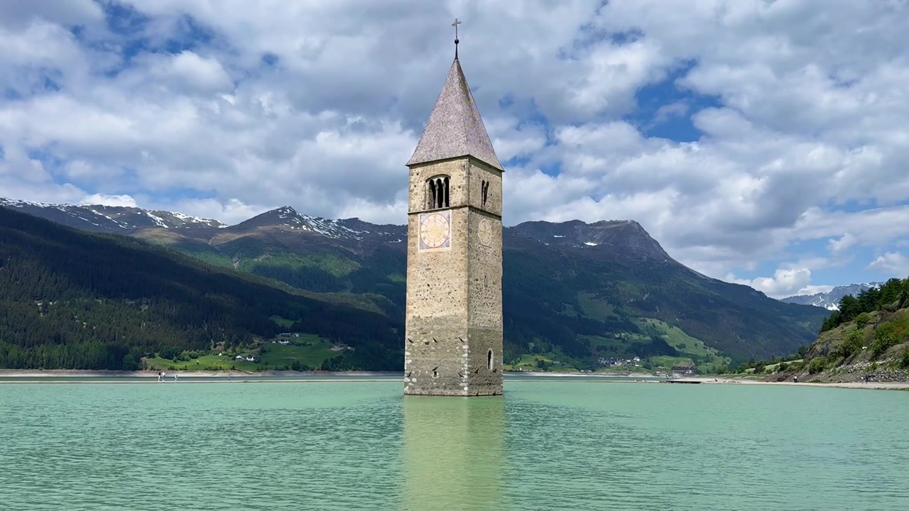 Vue fixe du clocher submergé de Curon dans le lac de Resia turquoise, sous des nuages en mouvement au début de l'été.