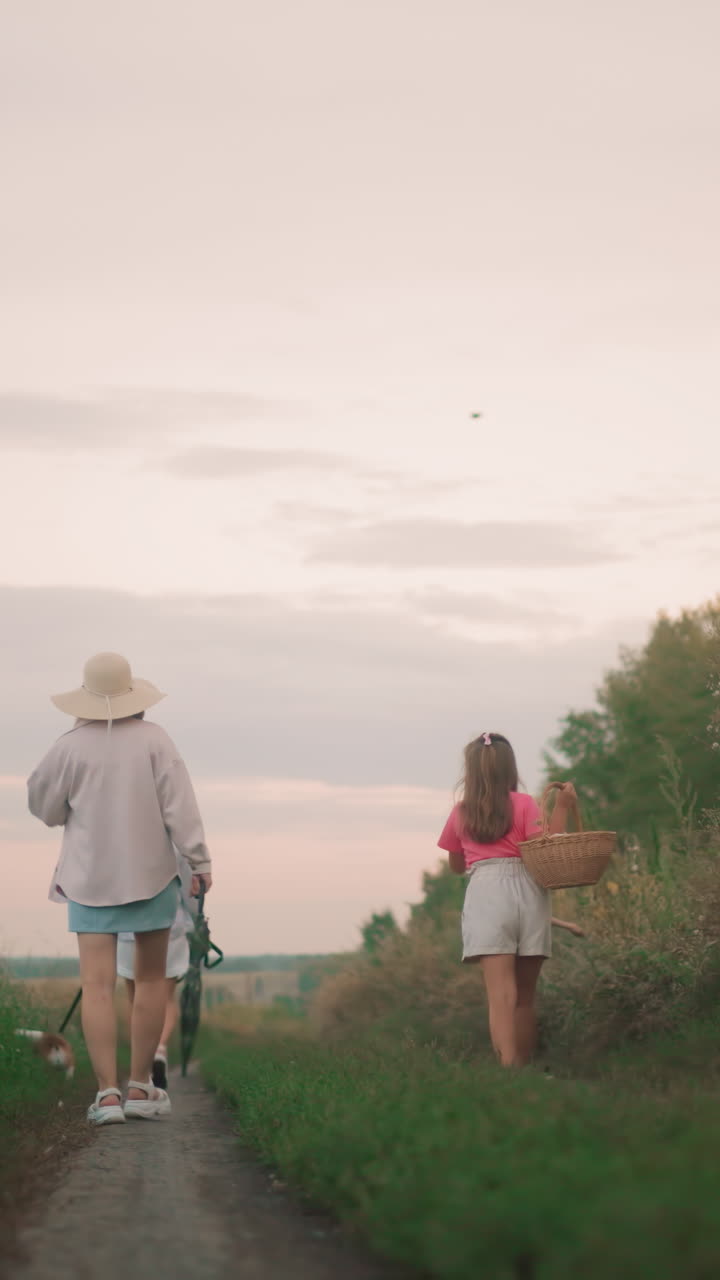 vista trasera de la familia caminando a casa a lo largo de un camino de tierra al atardecer, con un pájaro volando por encima, dos mujeres con sombreros caminando con un perro en una correa, mientras una niña joven lleva una canasta y un niño juega
