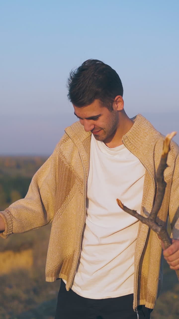 happy young man breaks stick for bonfire at comfortable camp on hill against pictorial meadows and blue evening sky