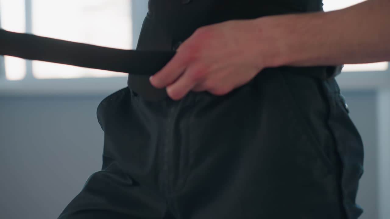 closeup waist of dancer removing belt from black pants in bright studio with tall windows behind highlighting contrast of smooth skin and textured fabric in elegant motion