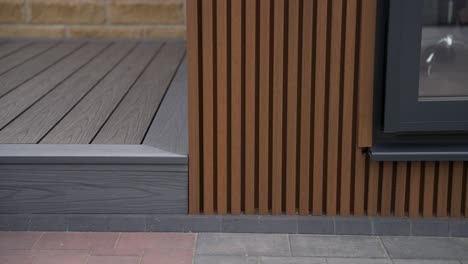 Close-up-on-new-exterior-garden-landscaping-with-luxury-grey-decking-running-next-to-perpendicular-brown-cladding-on-a-garden-room