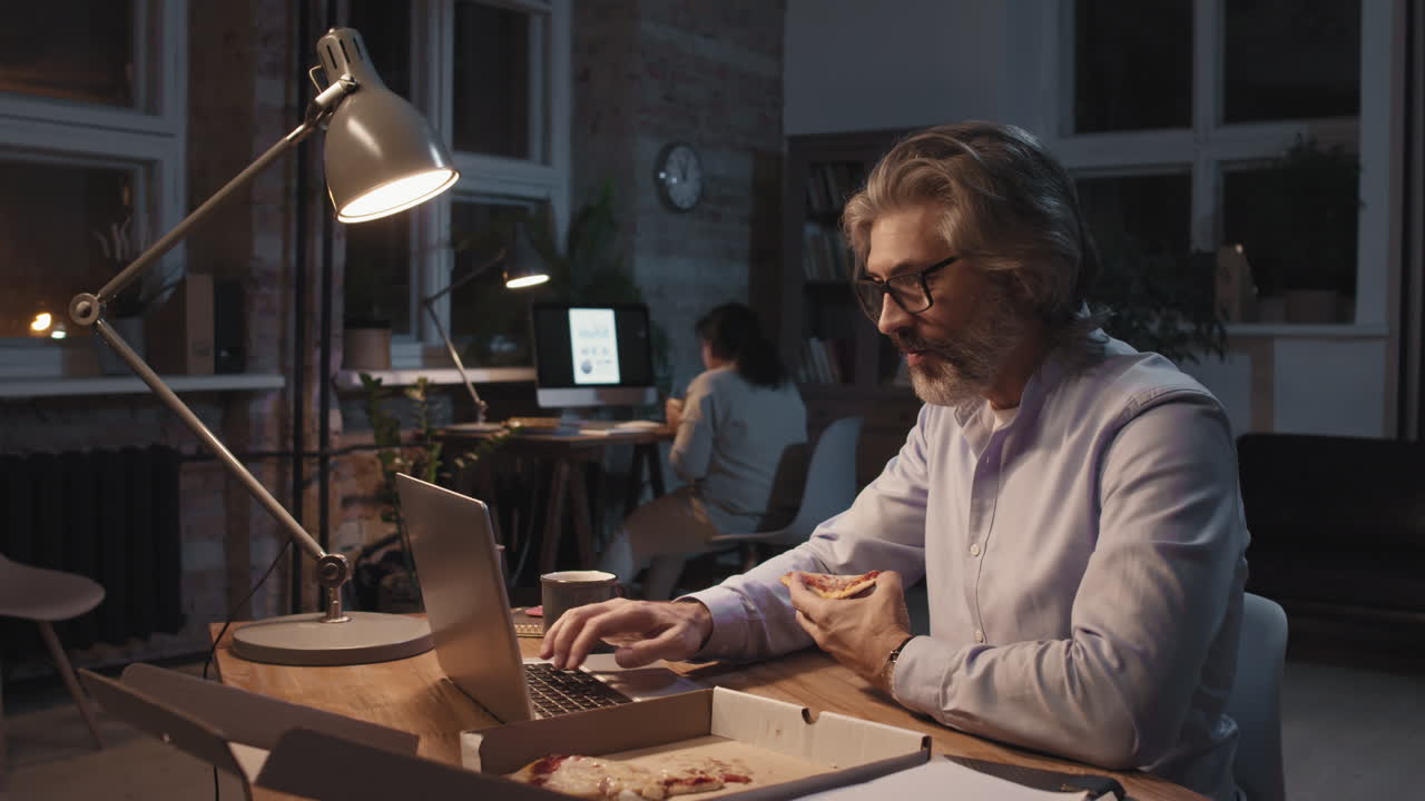 Man working late in the office, eating pizza and using a laptop