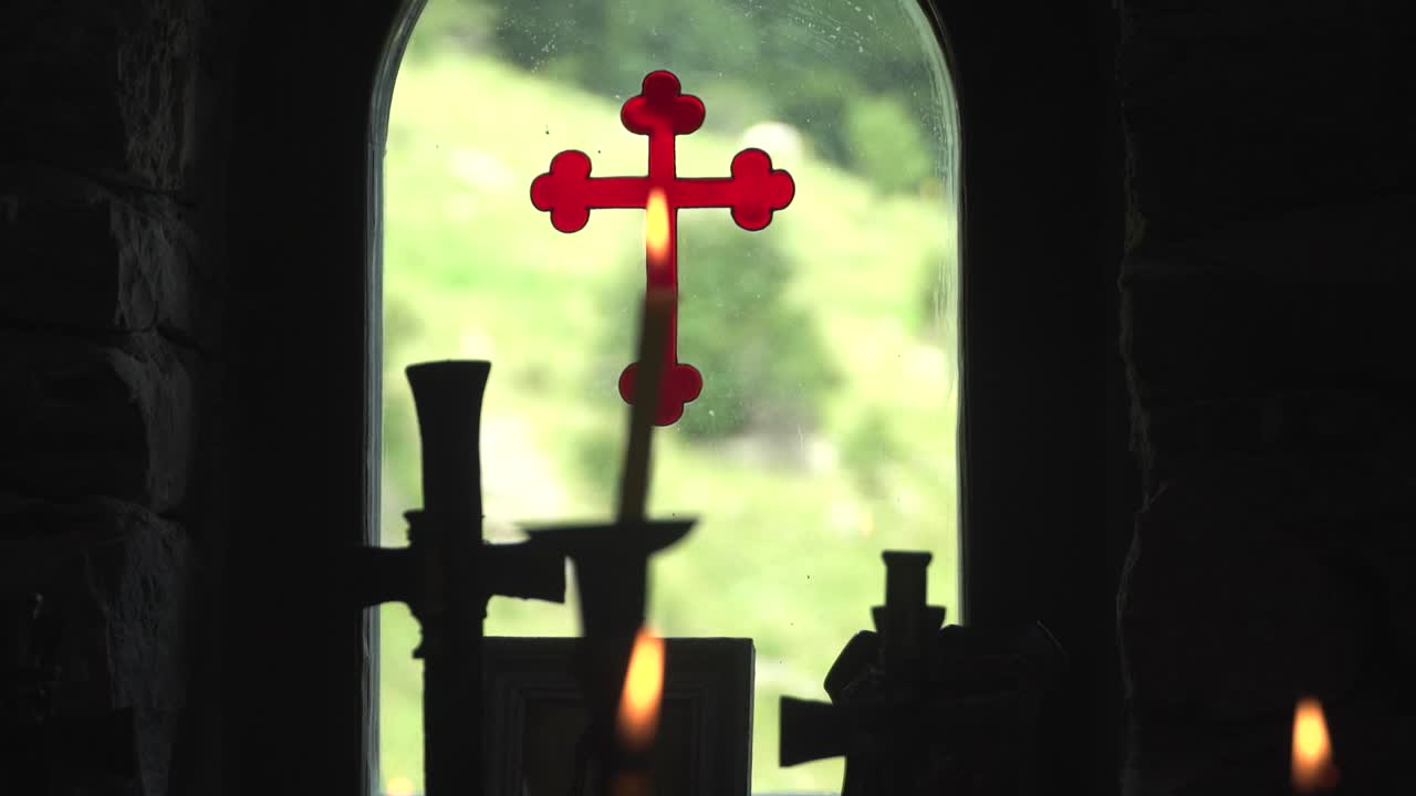 View from inside out through the window of a Bulgarian Orthodox Chapel. Burning candles against a background of a red cross painted on glass. Racking focus. Silhouette. Eastern Orthodox Church.