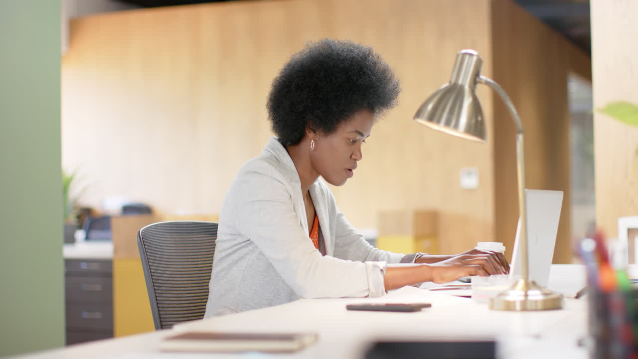 Happy african american casual businesswoman with afro using laptop in office, slow motion