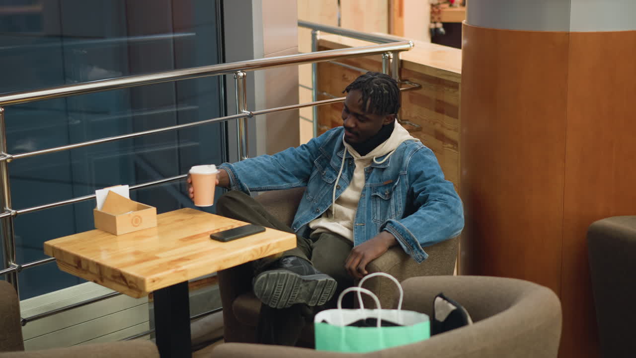 Guy in denim jacket and beige hoodie sits comfortably in armchair holding coffee cup at modern caf table, smartphone nearby, shopping bags on seat, relaxing moment after shopping in cozy public space