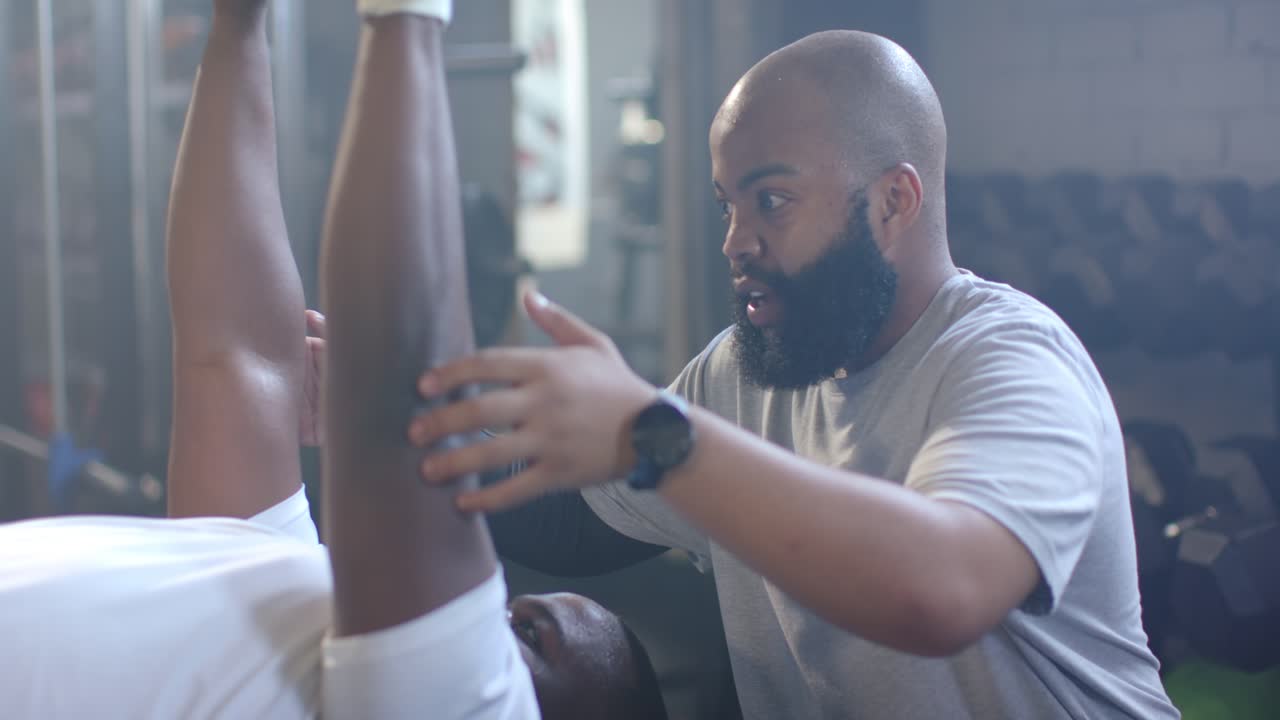 African American men trainer reaching spotting client pressing hex dumbbells on bench in gym safely