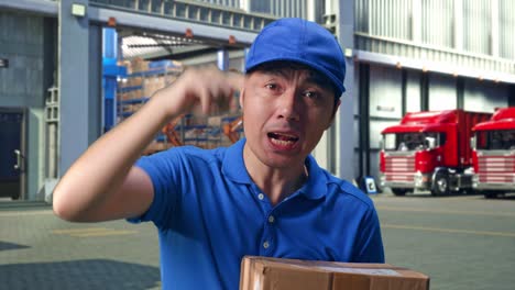close up of unsatisfied asian male courier in blue uniform shouting to camera while delivering a carton, outside of logistics distributions warehouse