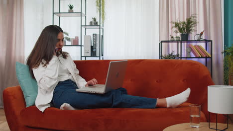 Young woman using laptop computer sitting on sofa working, online shopping from home office