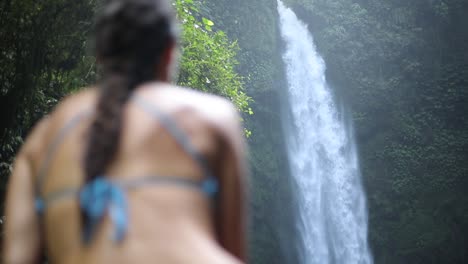 Slow-motion-panning-shot-of-a-girl-in-a-blue-bikini-sitting-in-front-of-a-gushing-NungNung-Waterfall-in-Bali,-Indonesia