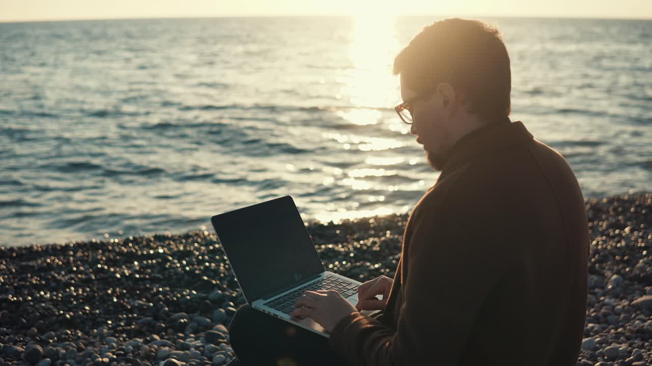 homem a trabalhar num portátil na praia ao pôr-do-sol