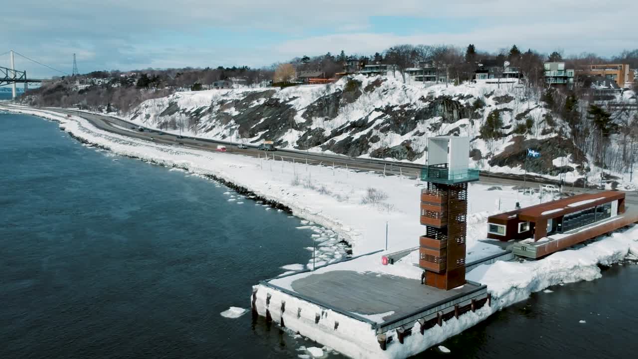 Modern observation tower rises above snowy pier near Riviere du Loup waterfront in Lower Saint Lawrence region as winter landscape meets cold river creating crisp scenic view from drone