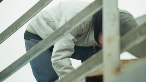 close-up of a young boy wearing a gray jacket, blue joggers, and a green beanie, focused on what he is doing near an iron railing