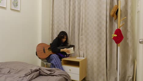 Wide-shot-of-an-Asian-woman-in-pajamas-writing-in-notebook-while-using-acoustic-guitar-in-her-bedroom