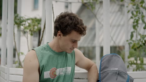 Young caucasian man sitting on a banch in the park and reading a booklet near his grey backpack arms and shoulders exposed