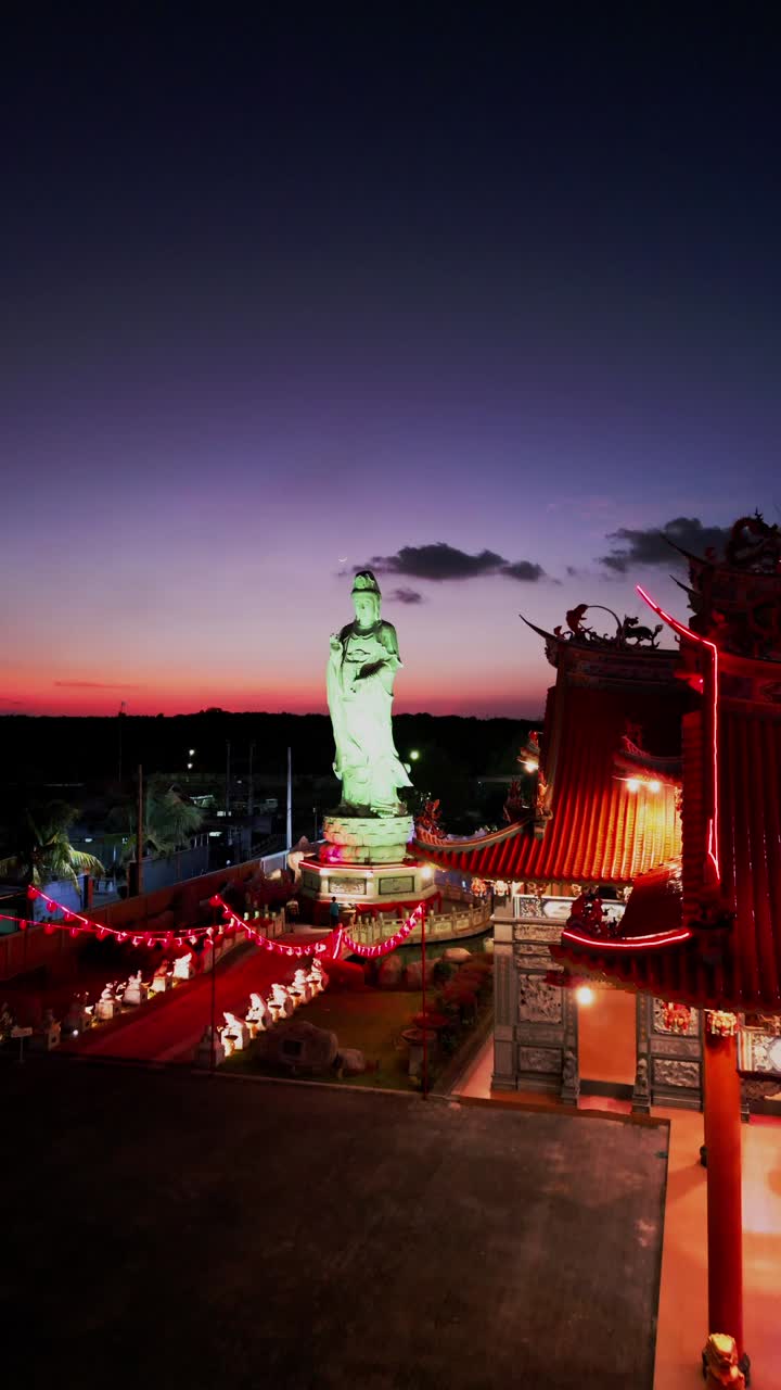 Vertical footage captures a Buddhist statue glowing in changing colors beside a traditional temple framed by a radiant sunset sky creating a serene blend of sacred architecture and nature