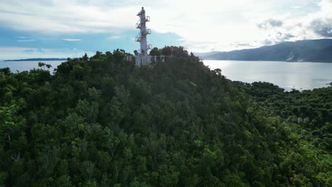 impresionante toma de drones en ascenso del faro de bote en la cima de montañas cubiertas de selva tropical con una impresionante bahía oceánica y nubes en el fondo