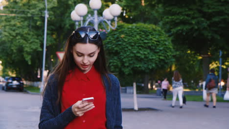 Atractiva-Mujer-Caminando-Por-La-Calle-Disfrutando-Del-Teléfono-Inteligente-Está-Escribiendo-Mira-Publicaciones