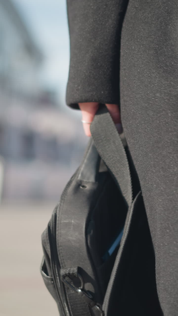 primer plano de una bolsa negra sostenida por una persona que lleva un elegante abrigo negro y botas de cuero, con la luz solar natural destacando los detalles elegantes de la ropa en un telón de fondo de calle urbana
