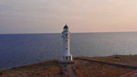 Amazing-lighthouse-stands-tall-on-the-side-of-a-cliff-during-majestic-sunset-reflecting-off-blue-ocean-waters,-drone-Aerial