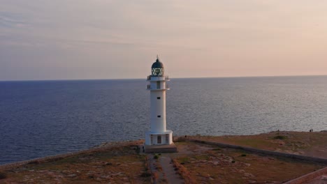 Asombroso-Faro-Se-Alza-En-La-Ladera-De-Un-Acantilado-Durante-La-Majestuosa-Puesta-De-Sol-Que-Se-Refleja-En-Las-Aguas-Azules-Del-Océano,-Antena-De-Drones