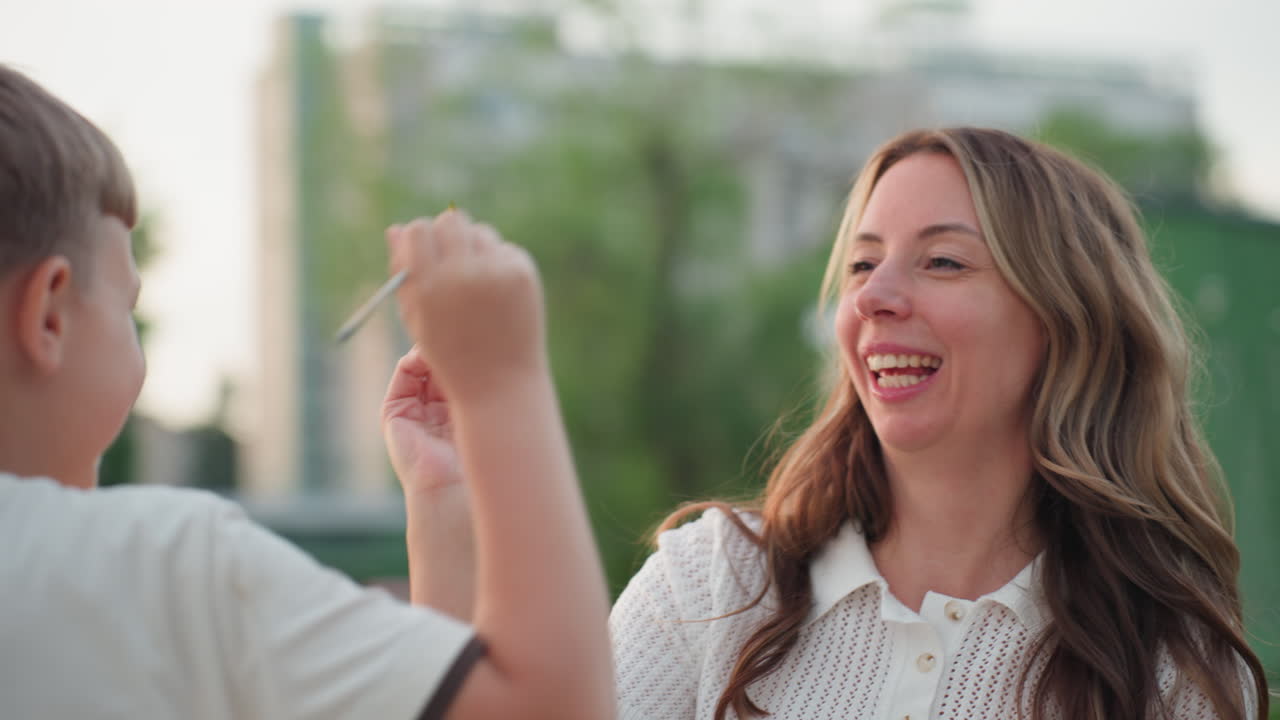 close up of child hand waving paint brush toward mom while seated on wooden dock by pool under sunset glow, capturing gentle playful shared art moment with water reflection and warm evening light