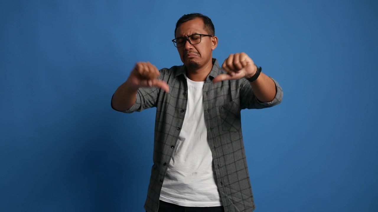 Asian adult man standing isolated on blue background, showing thumbs down with expression of displeasure, negative emotion, rejection or disagreement