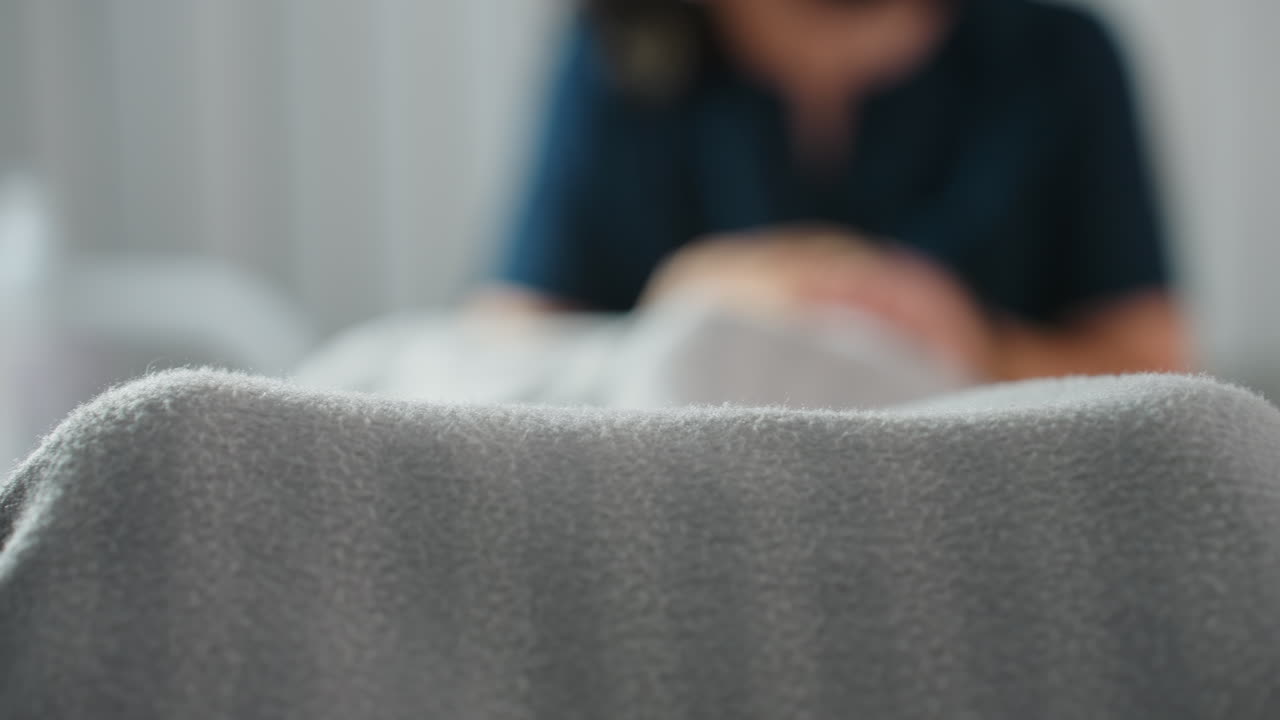 Female model covered in striped grey blanket lying on massage bed in bright spa room with faint blurred image of therapist massaging her during relaxing wellness session in serene environment