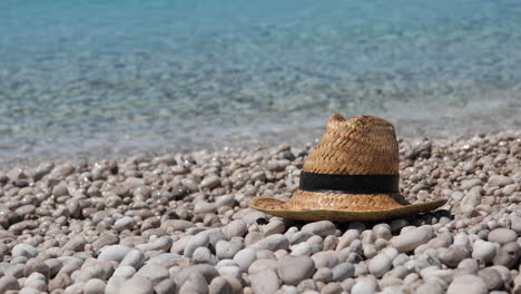 Sombrero-De-Paja-En-La-Playa-De-Guijarros-Con-Fondo-De-Agua-De-Mar-Cristalina,-Concepto-De-Vacaciones